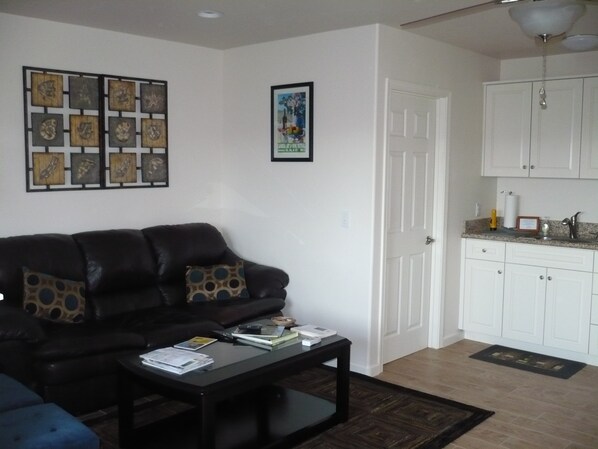A living room with a black leather sofa, a coffee table with books and papers, a framed picture on the wall, and a kitchen area with white cabinets and a countertop. - Located in the heart of Cambria. Stand alone upstairs one bedroom apartment. (Cambria)