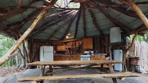 Una estructura rústica de madera con piso de piedra y techo de metal. En el interior, hay una zona de cocina con armarios de madera y un refrigerador.