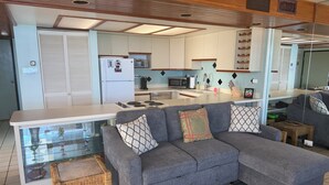 A modern kitchen with a grey sofa, a glass-top dining table, and a wicker bench. - Genuine & Affordable 1250 sqft Big Beach-front Corner Unit (WAIANAE)