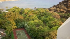 A tennis court surrounded by dense greenery and a view of the ocean. - Genuine & Affordable 1250 sqft Big Beach-front Corner Unit (WAIANAE)