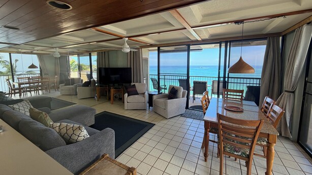 A spacious living area with a large TV, comfortable seating, a dining table, and a balcony with a view of the ocean. - Genuine & Affordable 1250 sqft Big Beach-front Corner Unit (WAIANAE)