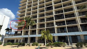A multi-story hotel with balconies, a beach in the foreground, and palm trees. - Genuine & Affordable 1250 sqft Big Beach-front Corner Unit (WAIANAE)