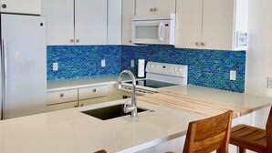 Una cocina moderna con armarios blancos, salpicadero de azulejos azules, microondas, estufa y un lavamanos con grifo. Hay tres sillas de madera en la isla.