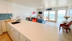 Una cocina moderna con una isla grande, un comedor con vistas al océano y una sala de estar con sofá y televisor.