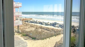 A beach view from a window, including a wooden deck, a building with a balcony, and a sandy beach with rocks. - Beach, Please: Best View of the Ocean with the Comforts of Home. 2bd/2ba (Carolina Beach)