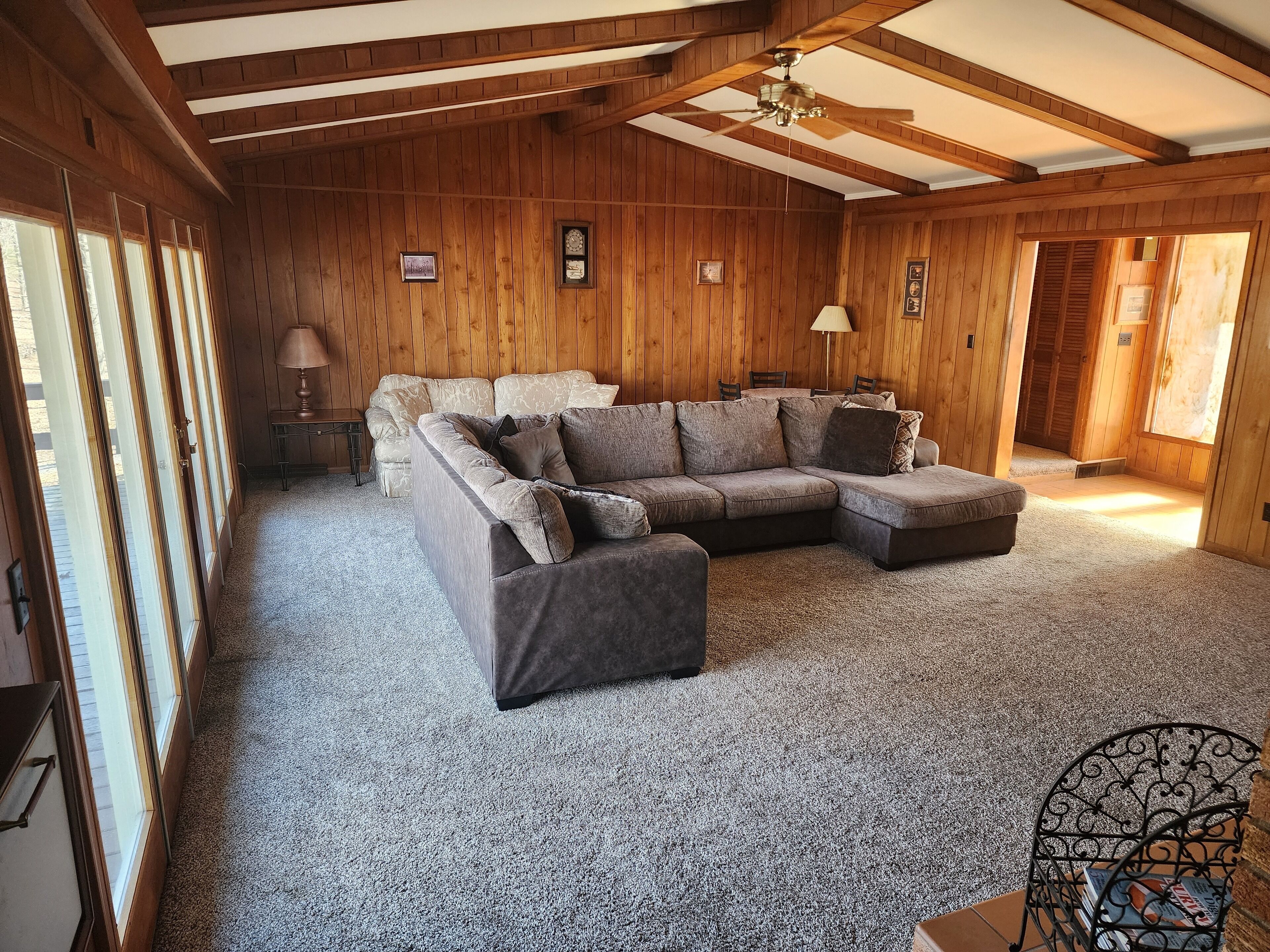 A cozy room with a sectional sofa, a small table, and a chair. There are wooden walls and a ceiling fan.