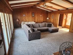 A cozy room with a sectional sofa, a small table, and a chair. There are wooden walls and a ceiling fan. - Kentucky Lake House on Jonathan Creek (Benton)