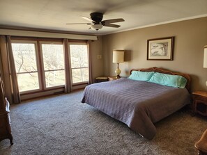 A bedroom with a large bed, a ceiling fan, a window with curtains, a bedside table with a lamp, and a framed picture on the wall. - Kentucky Lake House on Jonathan Creek (Benton)