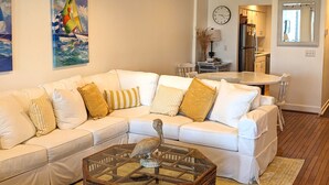 A modern living room with a sectional sofa, a glass coffee table with a pelican sculpture, a dining area with a round table and chairs, and a kitchen in the background. - Overlooks Pool & Ocean.  3 BR, 3 BA, Isle of Palms, SC. Near Charleston,S.C. (Isle of Palms)