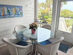 Un tavolo con piano in vetro, sedie in vimini, una pianta in vaso e un quadro con dei velieri sulla parete.