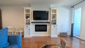 A living room with a flat-screen TV mounted above a fireplace, wooden flooring, and white cabinetry with shelves. - Overlooks Pool & Ocean.  3 BR, 3 BA, Isle of Palms, SC. Near Charleston,S.C. (Isle of Palms)