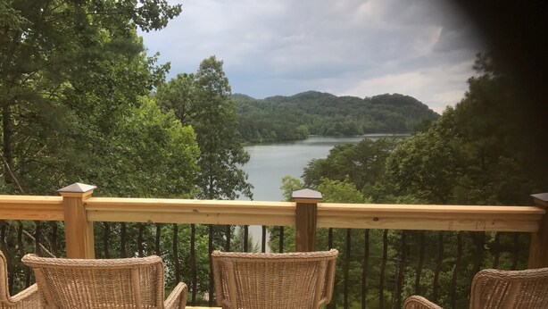 Una terraza de madera con sillas de mimbre que da a un lago y colinas boscosas.