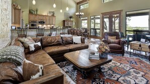 A spacious living room with a large leather sofa, a wooden coffee table, and a patterned rug. There is a fireplace, a dining area with chairs, and a kitchen in the background. - DREAM HOME w/ Amazing View! Movie Theater, Pool, Hot Tub, Game Room (San Marcos)