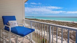 A balcony with a blue chair and a cushion, overlooking a beach with a pier. - Stay & Play | 🚲 Beachfront + Bikes, Kayaks, SUP & Pool Fun on Navarre Beach 🌴 (Navarre Beach)