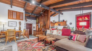 A cozy living room with a wooden ceiling, a sofa with patterned cushions, a coffee table, and a dining area with wicker furniture.