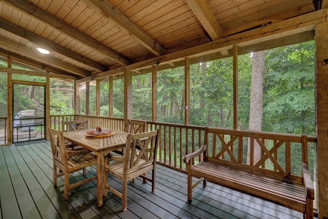 Une terrasse en bois avec une table et des bancs, entourée de grandes fenêtres donnant sur une forêt.