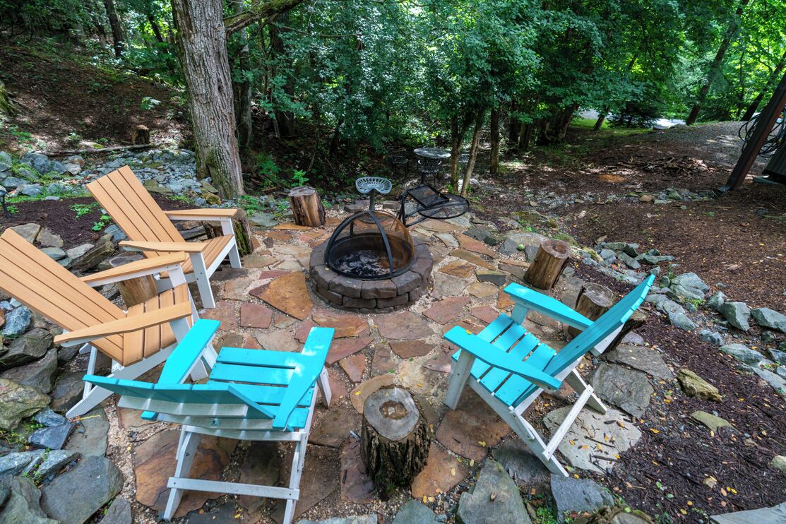 A stone fire pit surrounded by wooden Adirondack chairs in a forest setting.