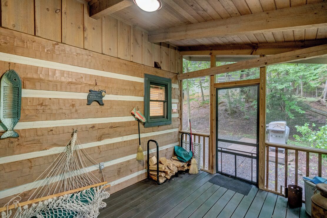 Interior de una cabaña con paredes de madera, una hamaca, chimenea y vistas al bosque.