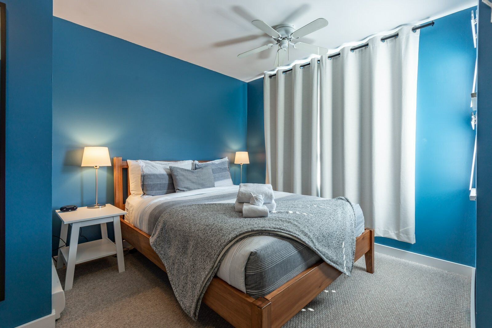 Un dormitorio con una cama, dos mesitas de noche, un ventilador de techo y una ventana con cortinas.