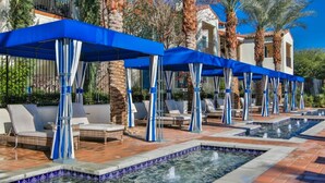 Un espace aménagé au bord de la piscine, avec des chaises longues et des parasols, entouré de palmiers et d’un bâtiment doté d’une piscine carrelée.