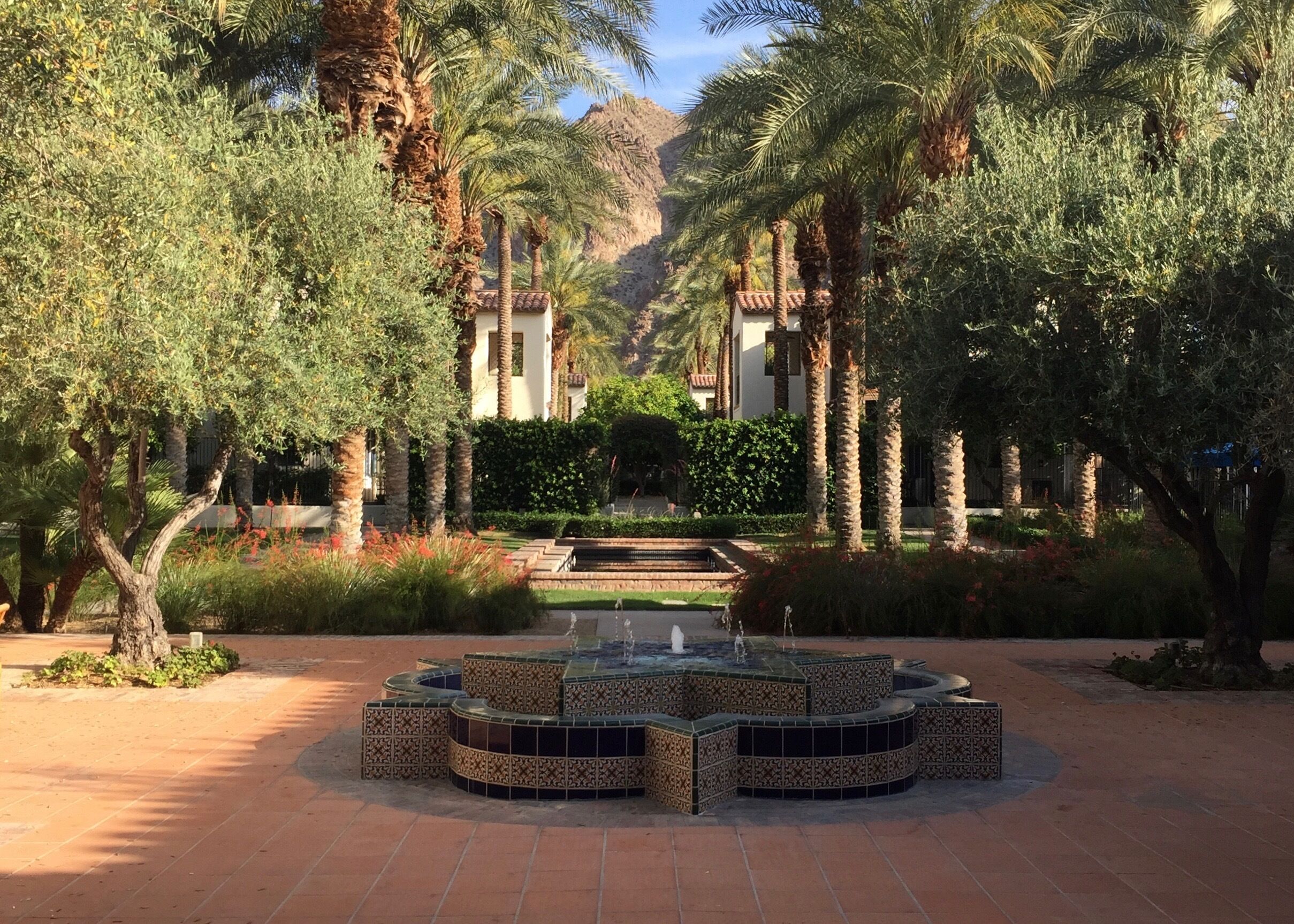 A courtyard with a patterned fountain, palm trees, and a building with a tiled roof in the background.