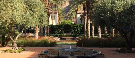 A courtyard with a patterned fountain, palm trees, and a building with a tiled roof in the background.