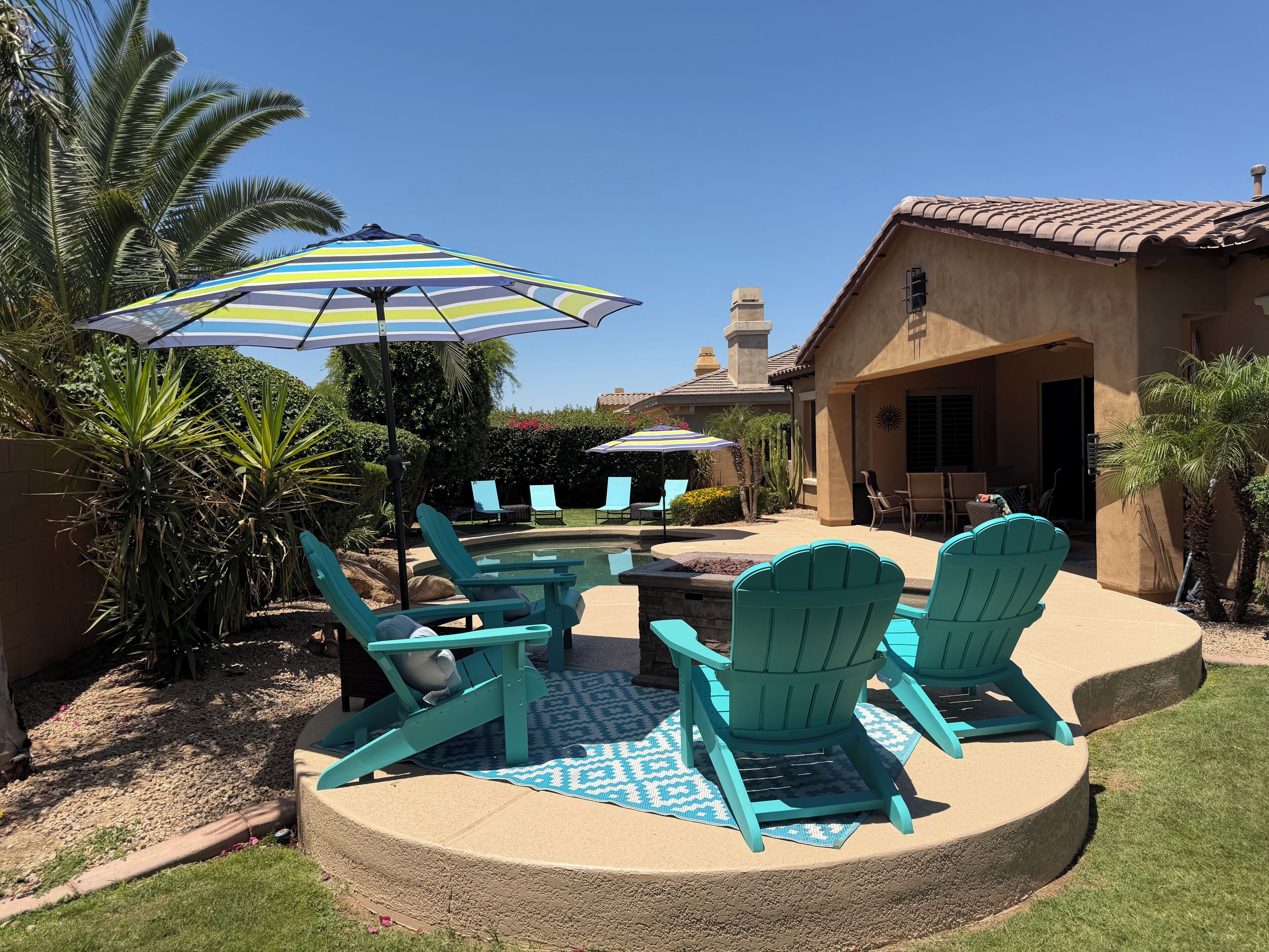 A pool area with turquoise Adirondack chairs, a patterned rug, and a striped umbrella.