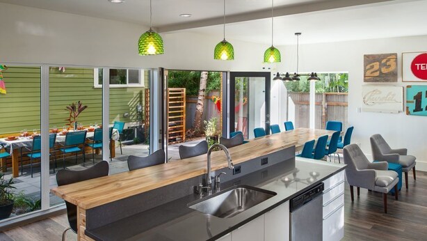 A modern kitchen with a central island, built-in dishwasher, and a dining area with blue chairs and a wooden table.