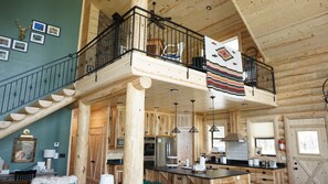 Cozy cabin-style interior with a high ceiling and exposed beams. Features a kitchen area with a central island and a mezzanine level with a balcony offering a view.