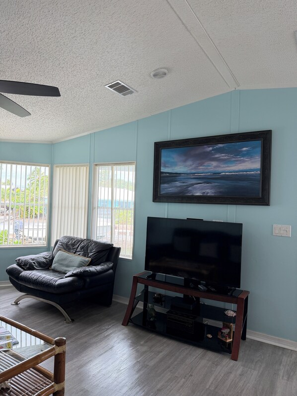A living room with a black leather sofa, a glass coffee table, a flat-screen TV on a wooden stand, and a painting of a seascape on the wall.