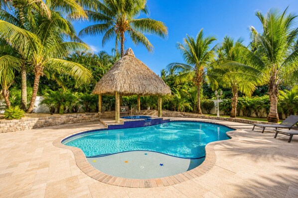 A swimming pool with a thatched roof structure, surrounded by palm trees and lounge chairs.