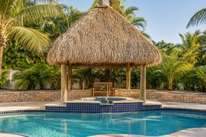 A swimming pool with a thatched gazebo, surrounded by palm trees and a brick wall.