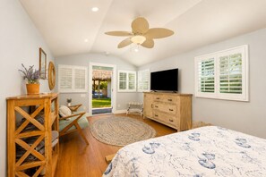 A bedroom with a bed, a TV, a wooden dresser, a ceiling fan, and a view of the outdoors.