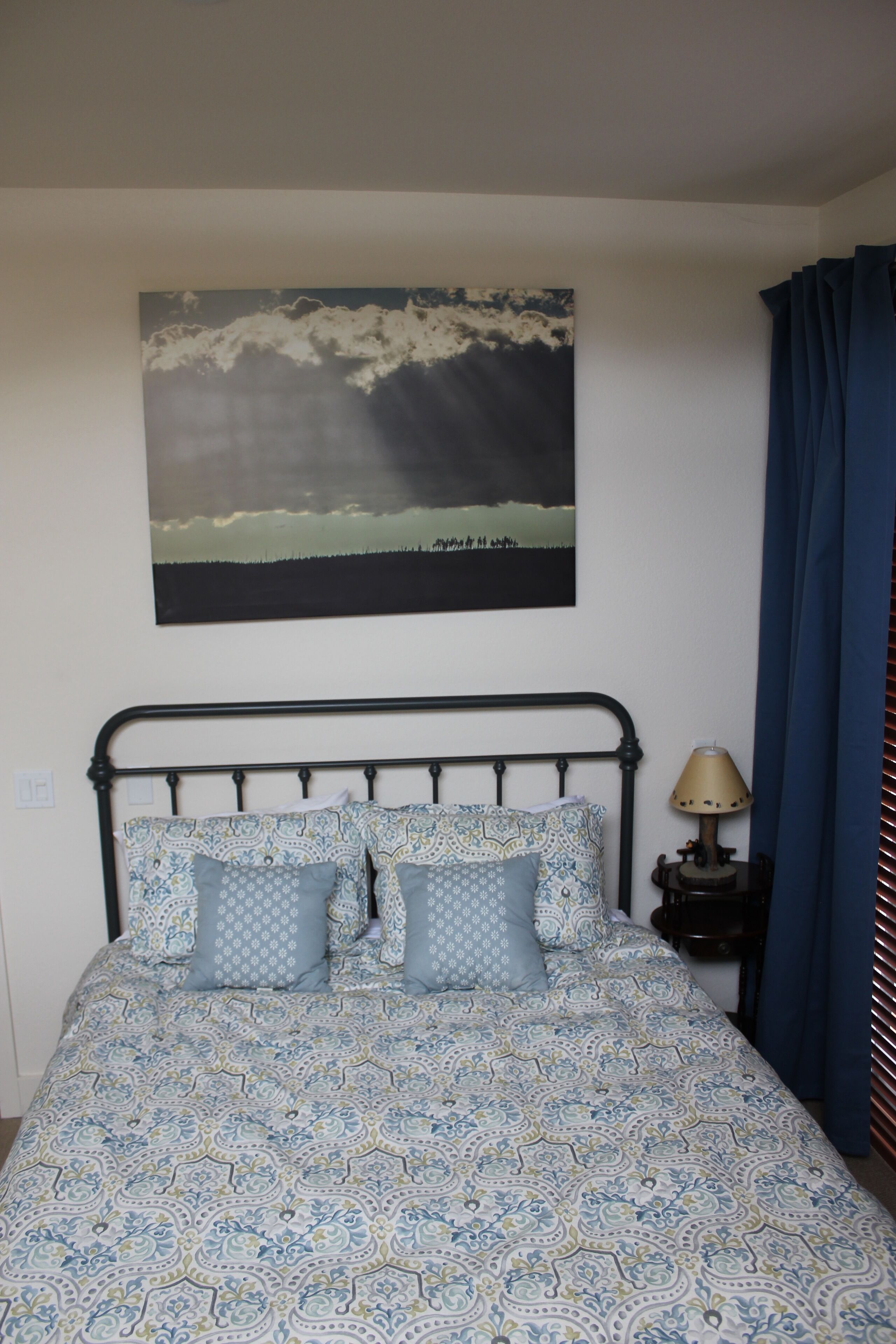 Un dormitorio con una colcha estampada, un cabecero, una lámpara de noche y una obra de arte en la pared que representa un paisaje.