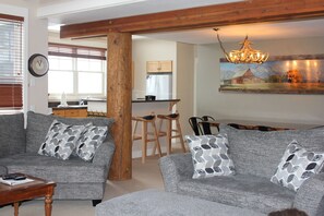 A living room with a grey sofa set, wooden beams, a chandelier, and a kitchen in the background. - Truly Ski In/Ski Out Condo!  Spacious- Sleeps 11!  Great Mountain Views! (Big Sky)