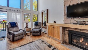 A spacious living room with two grey armchairs, a wooden coffee table, a fireplace, a large TV, and a stone wall. - Seconds to mall and slope, 2 hot tubs,1 pool, small gym, fireplaces (Snowmass Village)