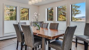 A dining room with a wooden table, six upholstered chairs, a chandelier, and large windows with a view of trees and hills. - Seconds to mall and slope, 2 hot tubs,1 pool, small gym, fireplaces (Snowmass Village)