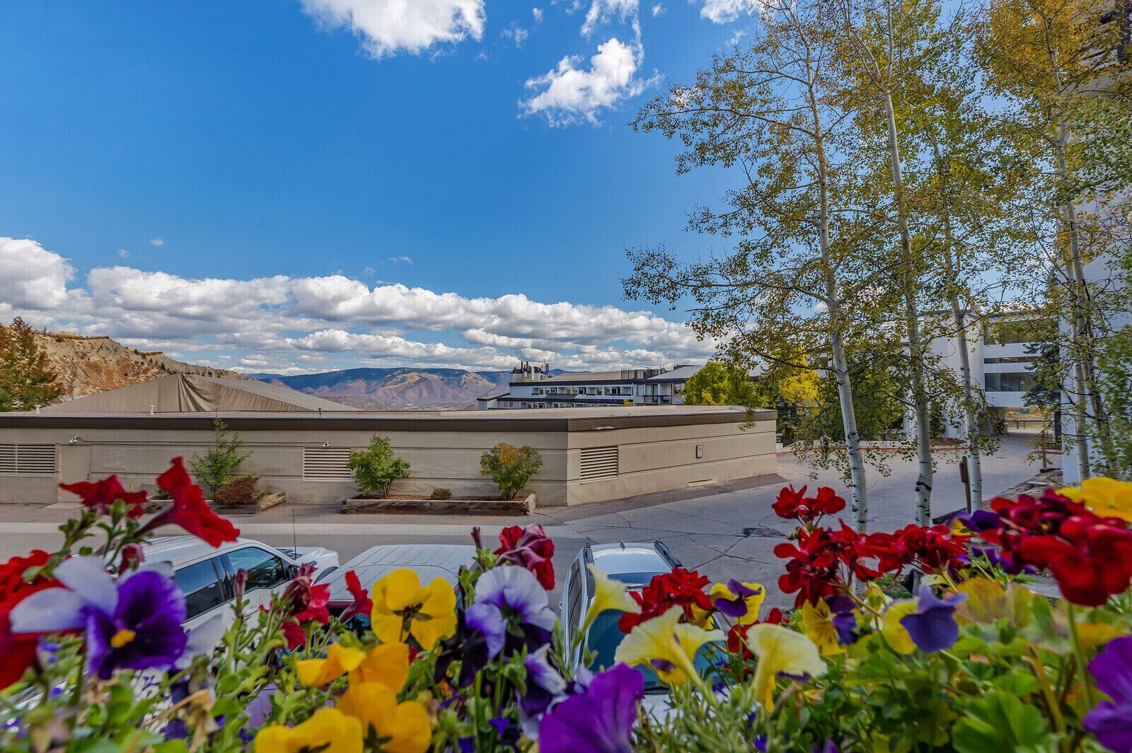 Un parking agrémenté de fleurs aux couleurs vives au premier plan, un bâtiment doté d’un toit plat et des montagnes en arrière-plan.