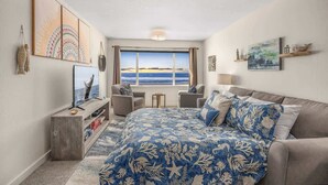A bedroom with a large bed, a TV, a desk, and a window with a view of the sea.