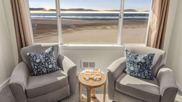 A room with two grey armchairs, a small wooden table, and a view of the beach through large windows.