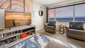 A living room with a large TV, bookshelves, two armchairs, a coffee table, and a view of the ocean.
