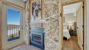 Une cheminée en pierre agrémentée d’une pancarte accueillante, une porte donnant sur une chambre, et une vue sur les montagnes à travers une porte vitrée.