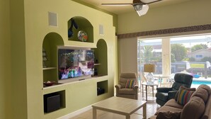Ein Wohnzimmer mit Flachbildfernseher an der Wand, einer Couch, einem Couchtisch und Blick auf einen Pool im Freien.