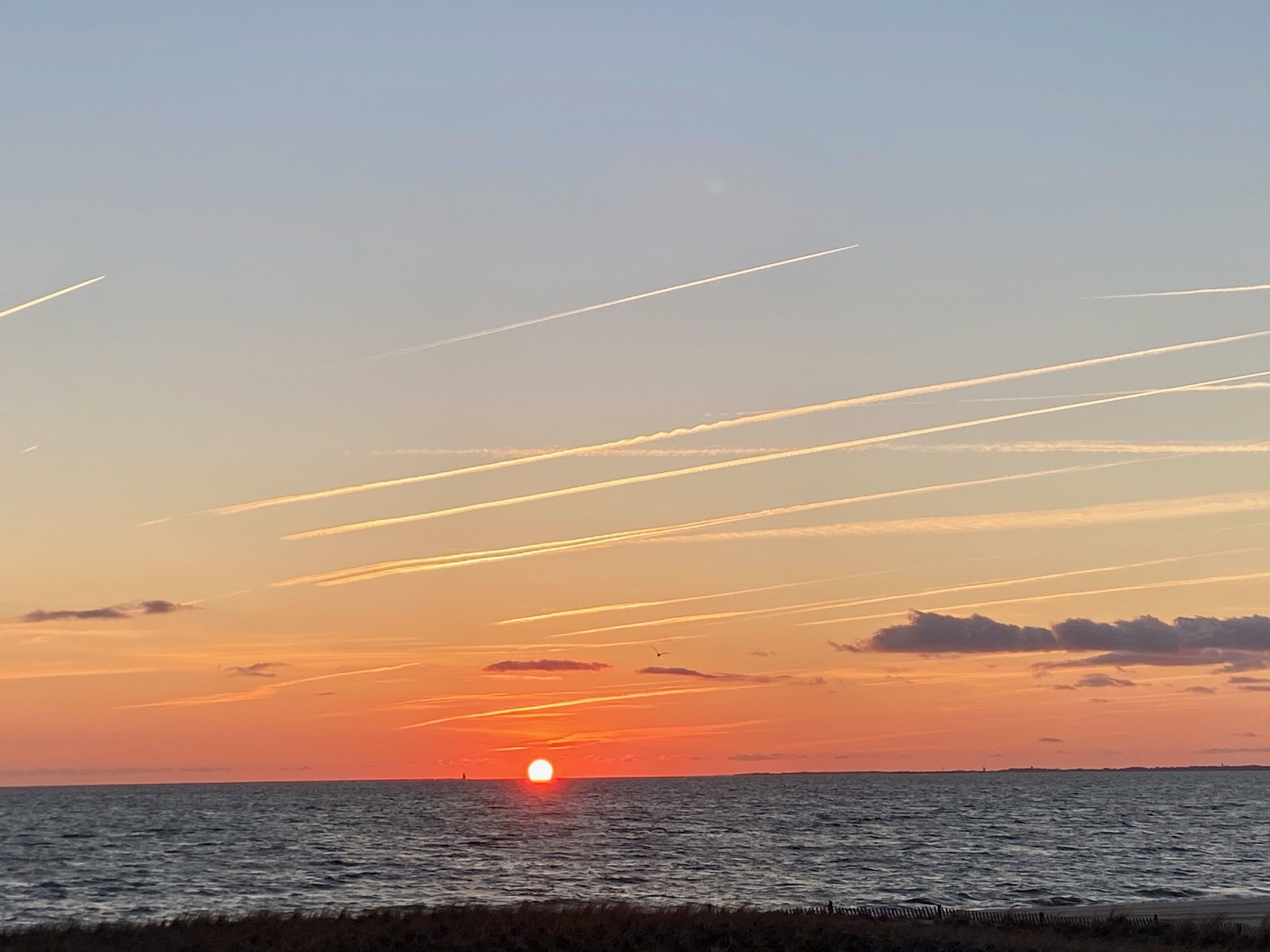 Auringonlasku valtameren yllä, taivaalla näkyy lentokoneiden jättämiä kondenssijälkiä.