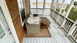 Een overdekt terras met een houten vloer, een zithoek met kussens, een bank en een kleine tafel, en uitzicht op het strand door grote ramen.