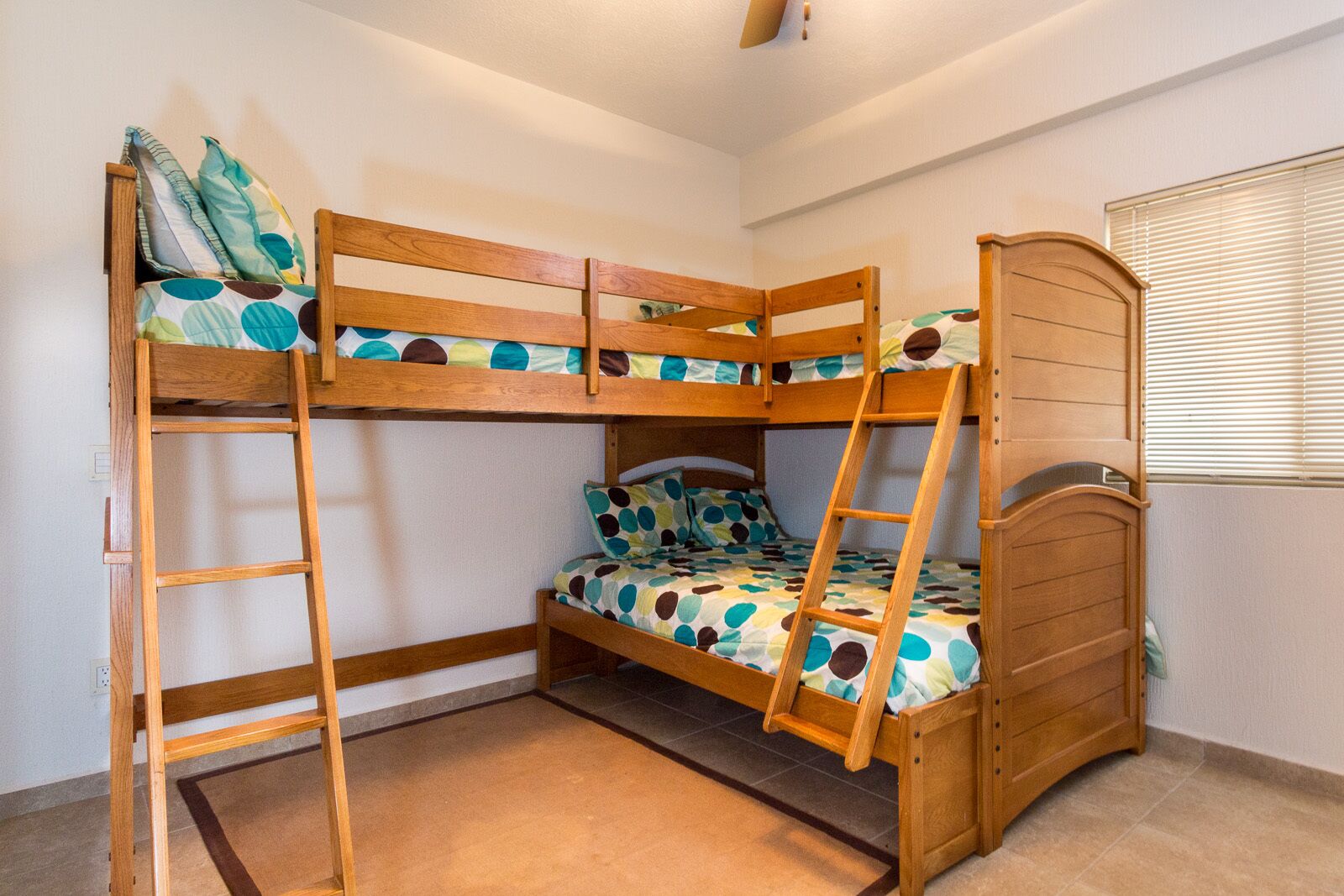 Une chambre avec un lit superposé, une échelle en bois, une fenêtre avec des stores et un sol carrelé.
