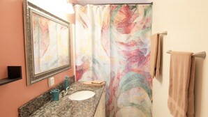 Une salle de bain avec un comptoir en granit, un lavabo, un miroir, un rideau de douche orné d’un motif abstrait et coloré, et un porte-serviettes avec une serviette.