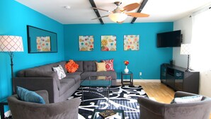 Un salon avec un canapé d’angle gris, une table basse et un tapis à motifs. On y trouve deux fauteuils, un téléviseur et un ventilateur de plafond.