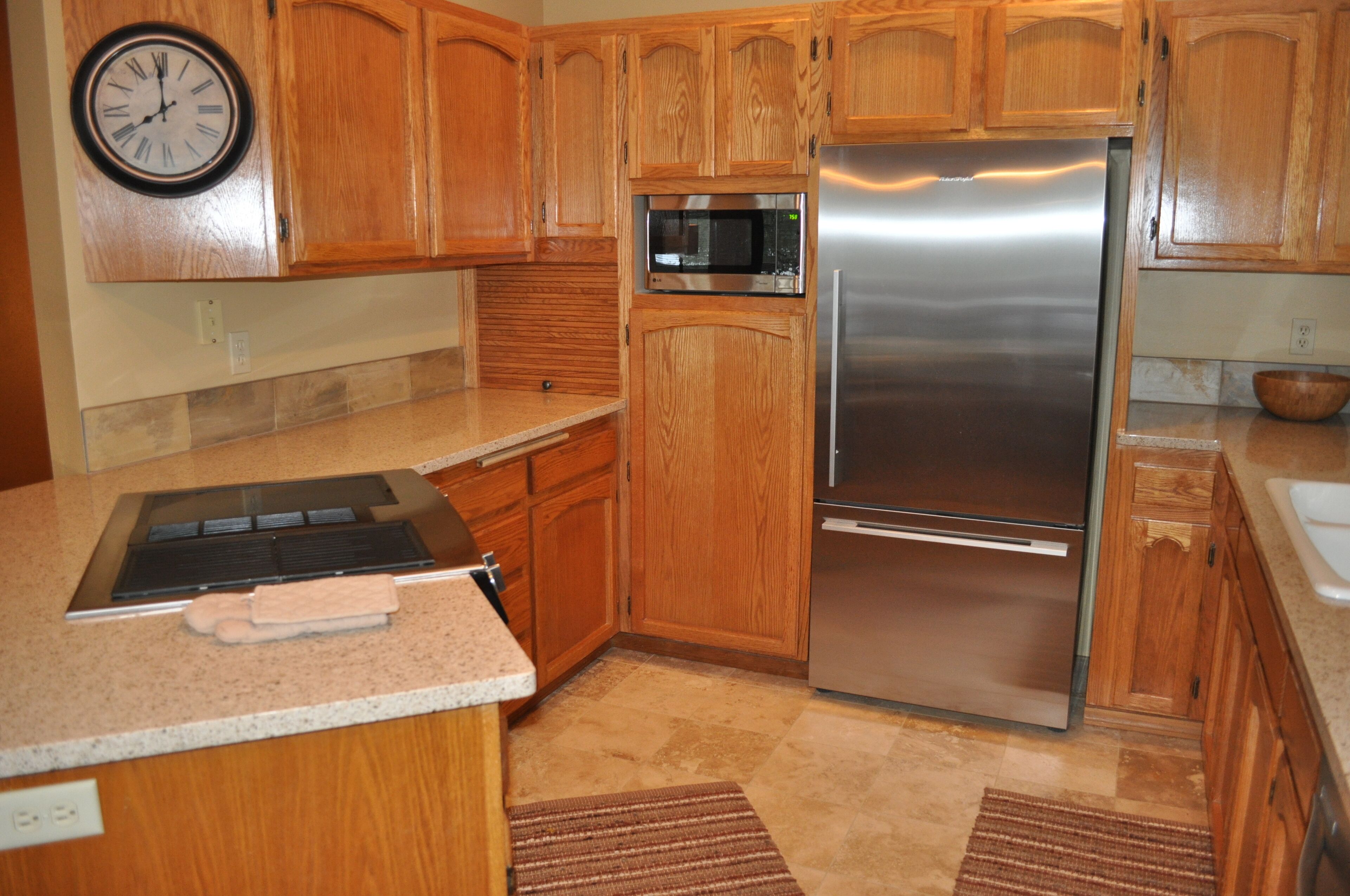 A kitchen with wooden cabinets, a stainless steel refrigerator, a microwave, and a clock on the wall.