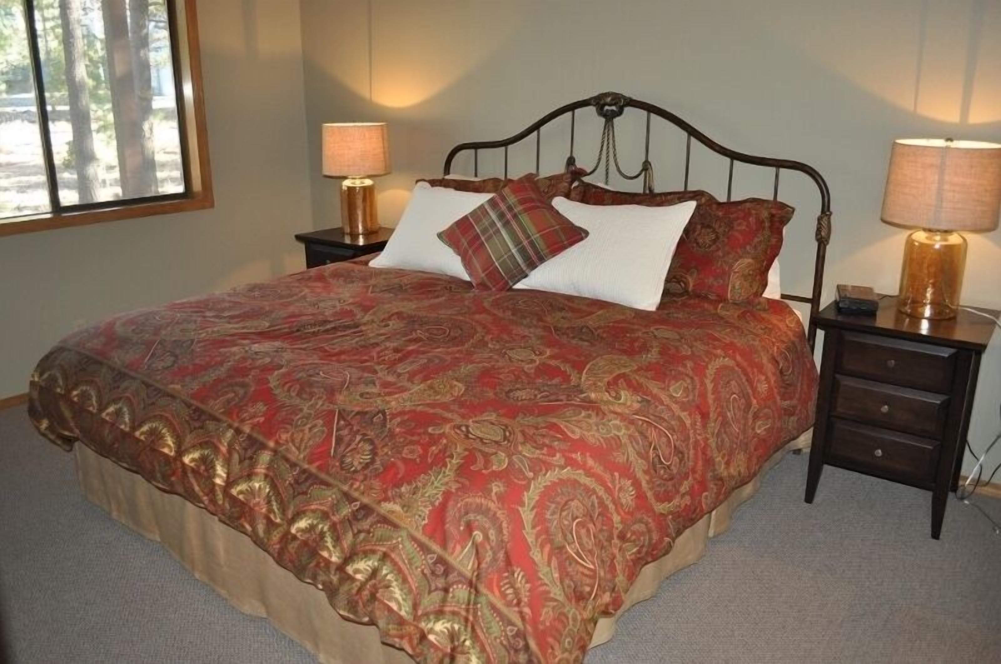 A bed with a red and gold patterned bedspread, two bedside tables with lamps, and a window with a view of trees.